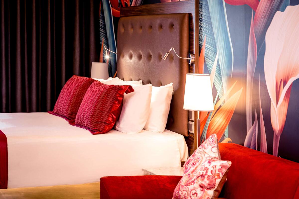 Une chambre d’hôtel avec un grand lit, des oreillers rouges et blancs, un fauteuil rouge et un mur orné d’une fresque florale.
