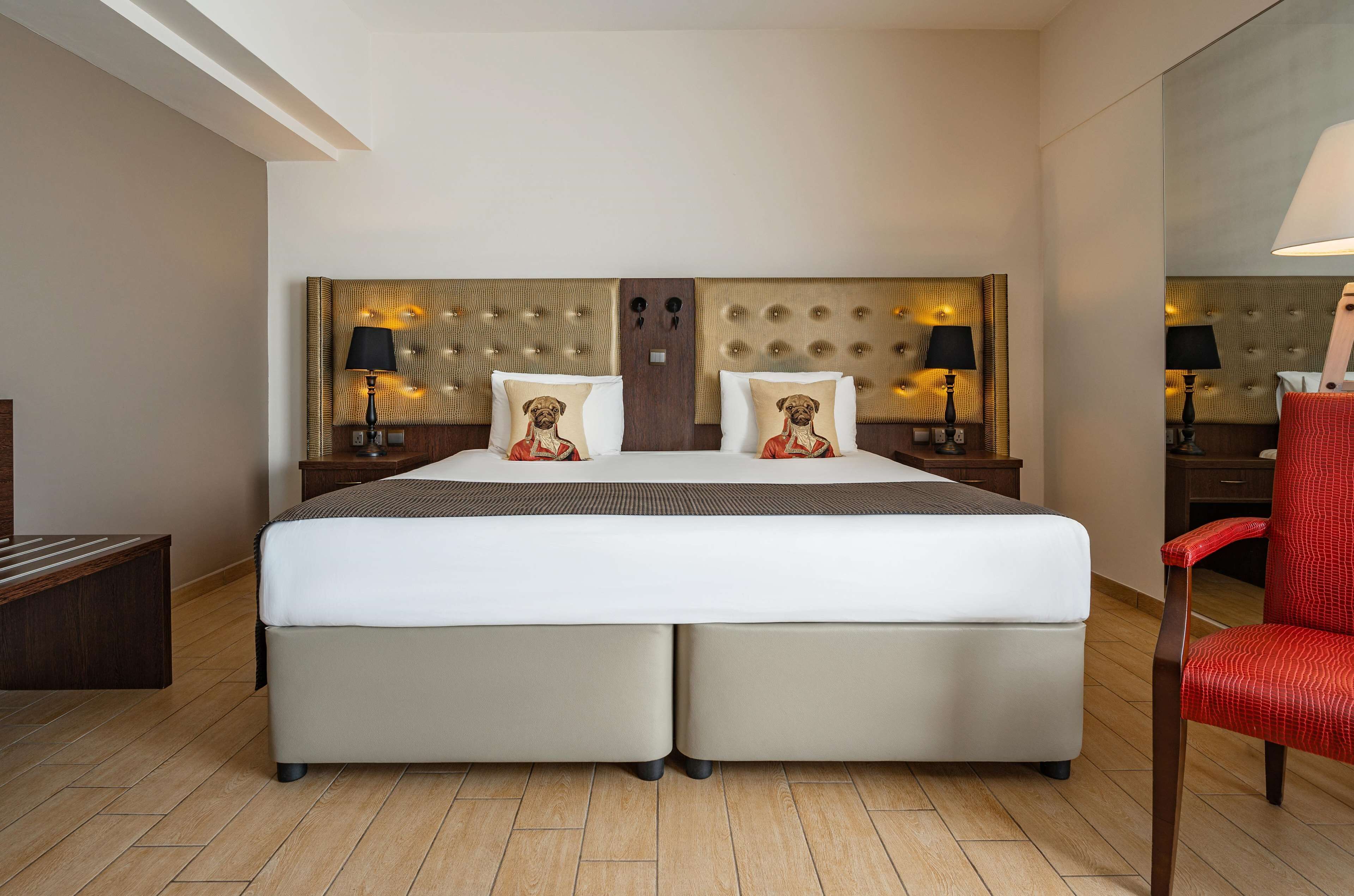 A double bed with a tufted headboard, two bedside tables with lamps, a red chair, and a wooden bench.