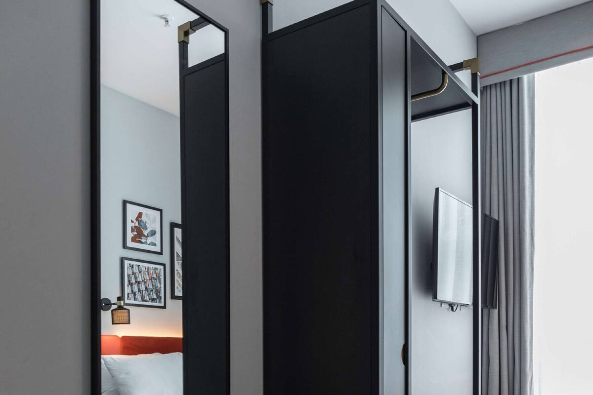 Moderni hotellihuone, jossa on suuri peili, valkoisilla lakanoiden peitetty sänky, pieni jalkarahi ja seinälle asennettu televisio.
