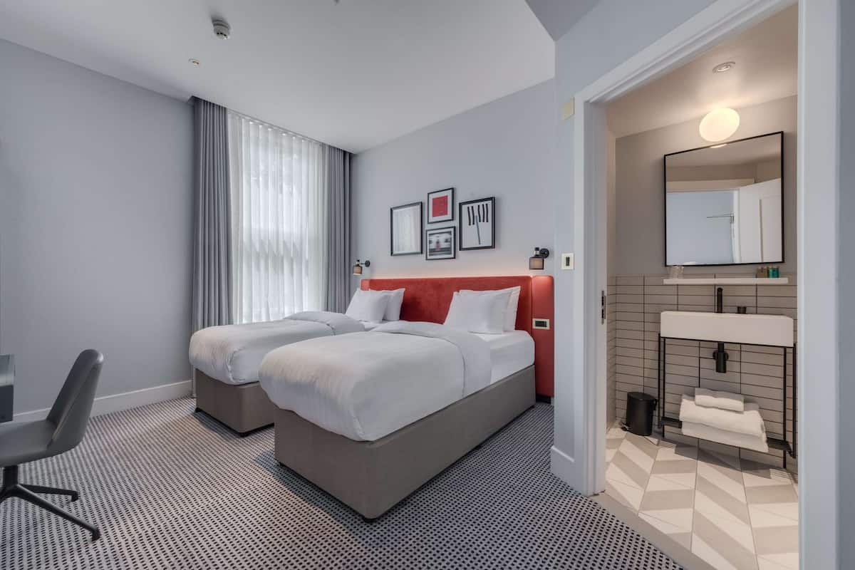 Hotellihuone, jossa on kaksi sänkyä, työpöytä, tuoli, peilillä varustettu kylpyhuone ja kuviollinen lattia.