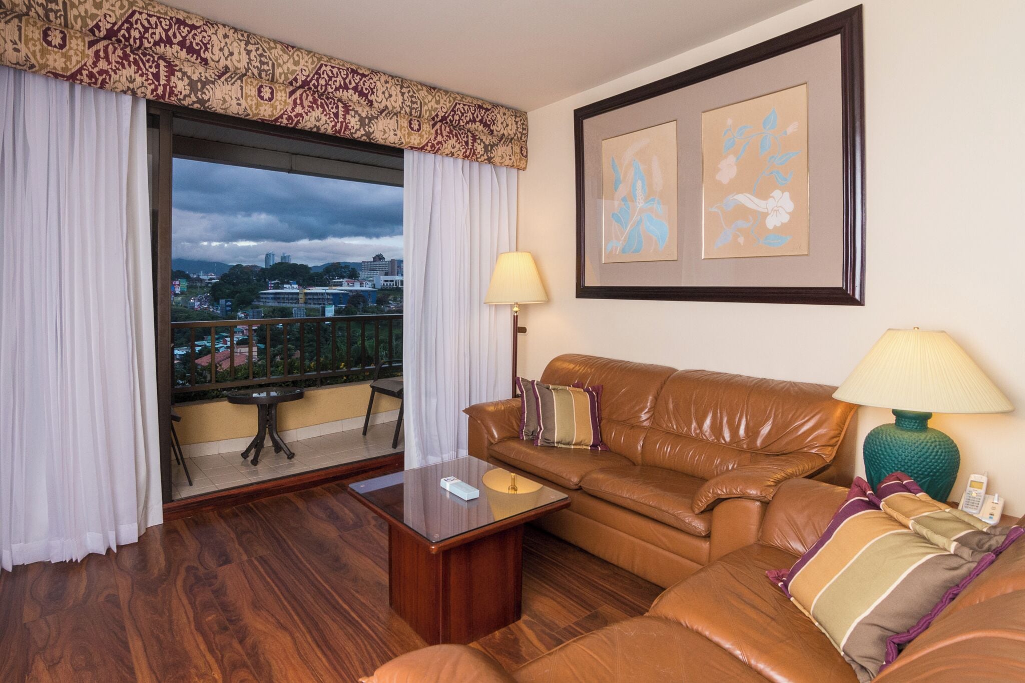 Una habitación de hotel con sofá de cuero, una mesita, un balcón con vistas a la ciudad y dos cuadros enmarcados en la pared.