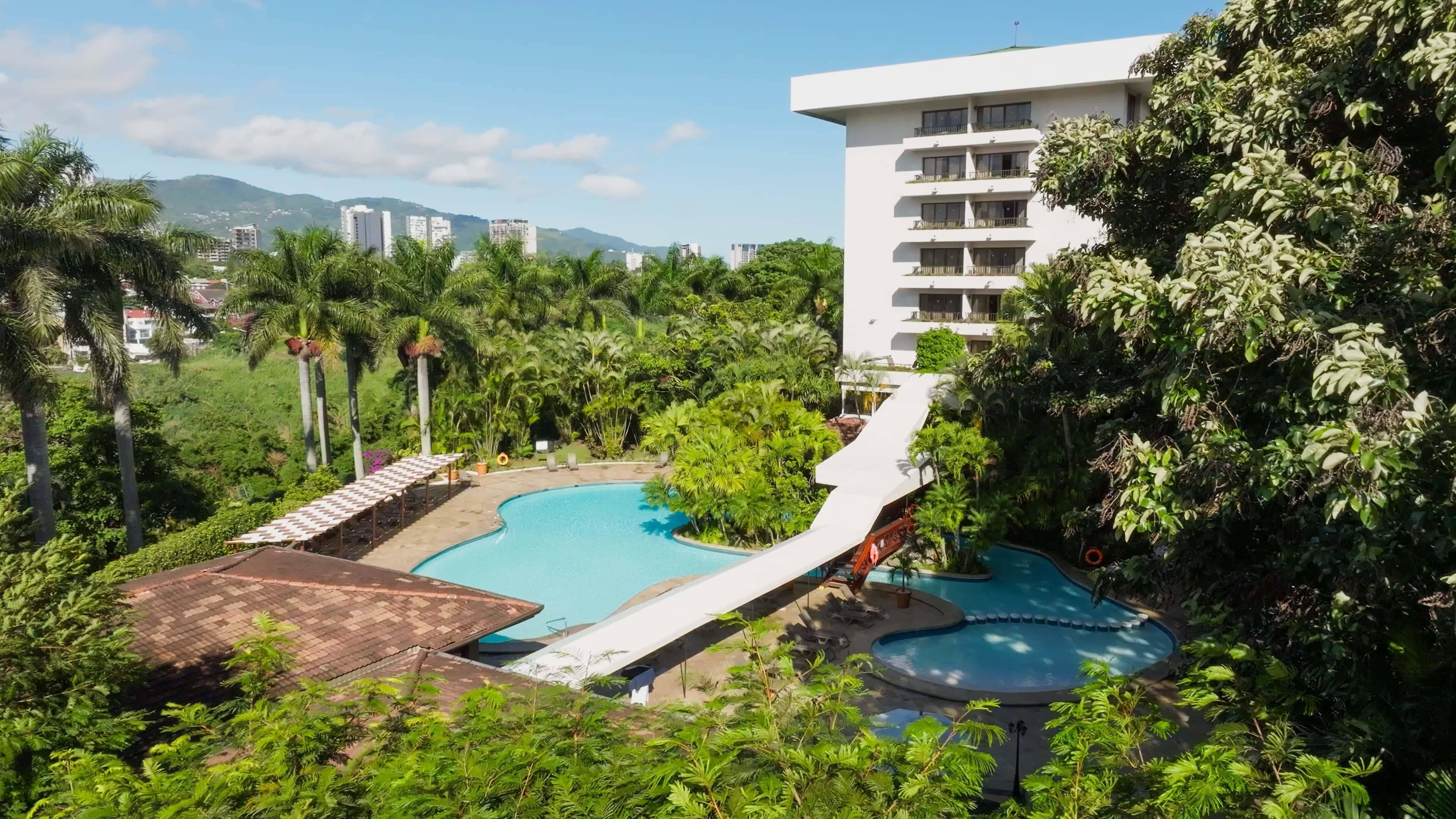 A resort with a large pool, lush greenery, and a view of distant buildings.