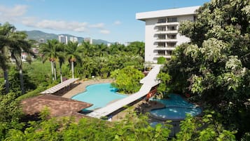 A resort with a large pool, lush greenery, and a view of distant buildings.