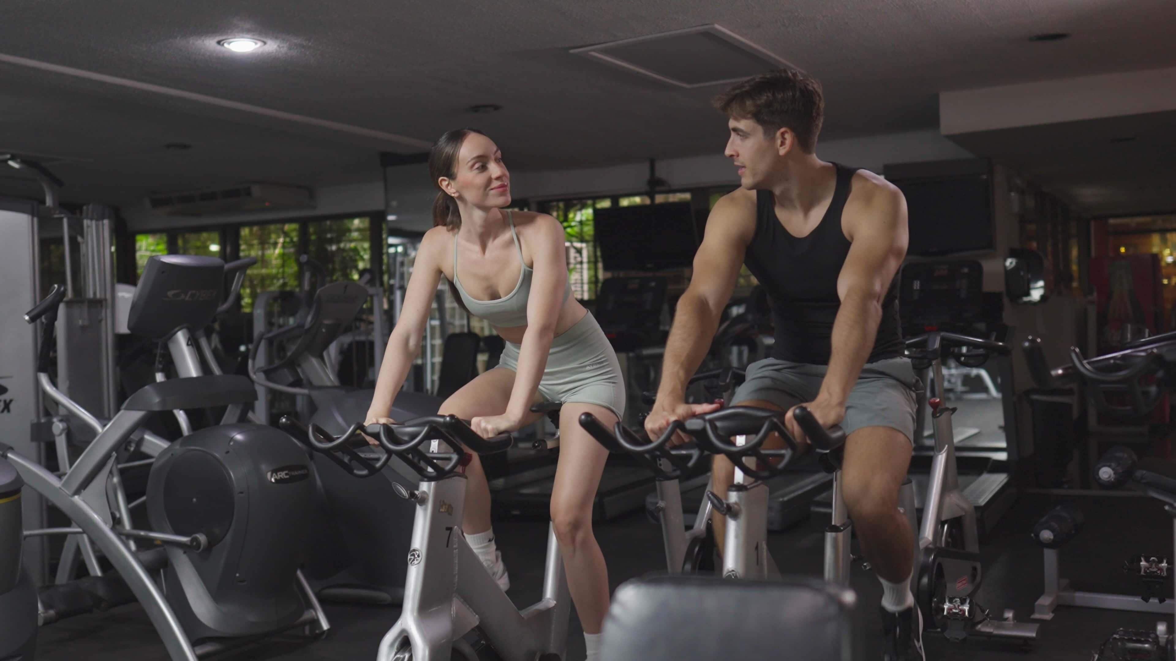 Duas pessoas pedalando em bicicletas ergométricas numa academia com diversos equipamentos de exercício.