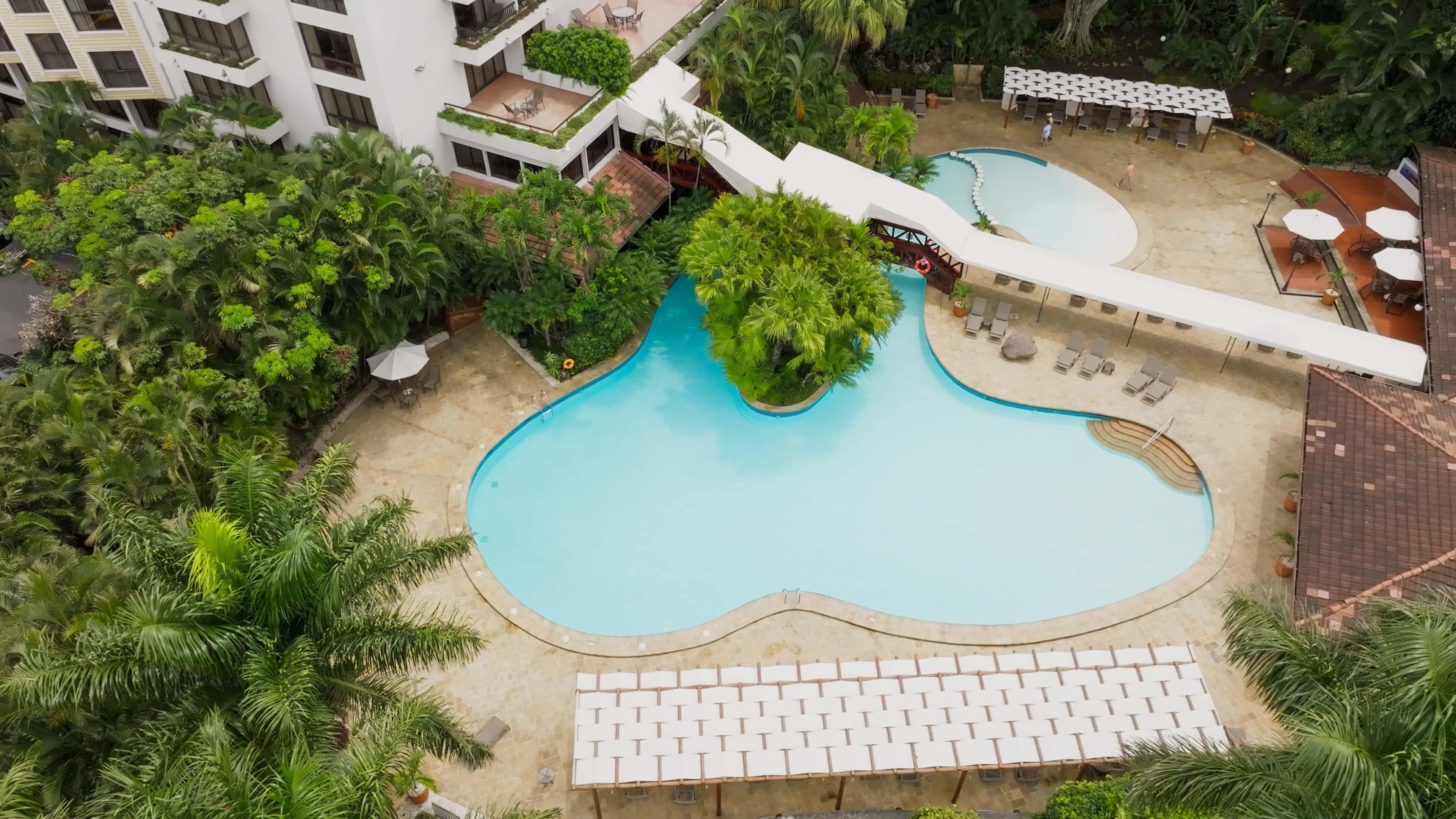 An aerial view of a hotel with a central swimming pool surrounded by lush greenery, outdoor seating areas, and a modern building structure.