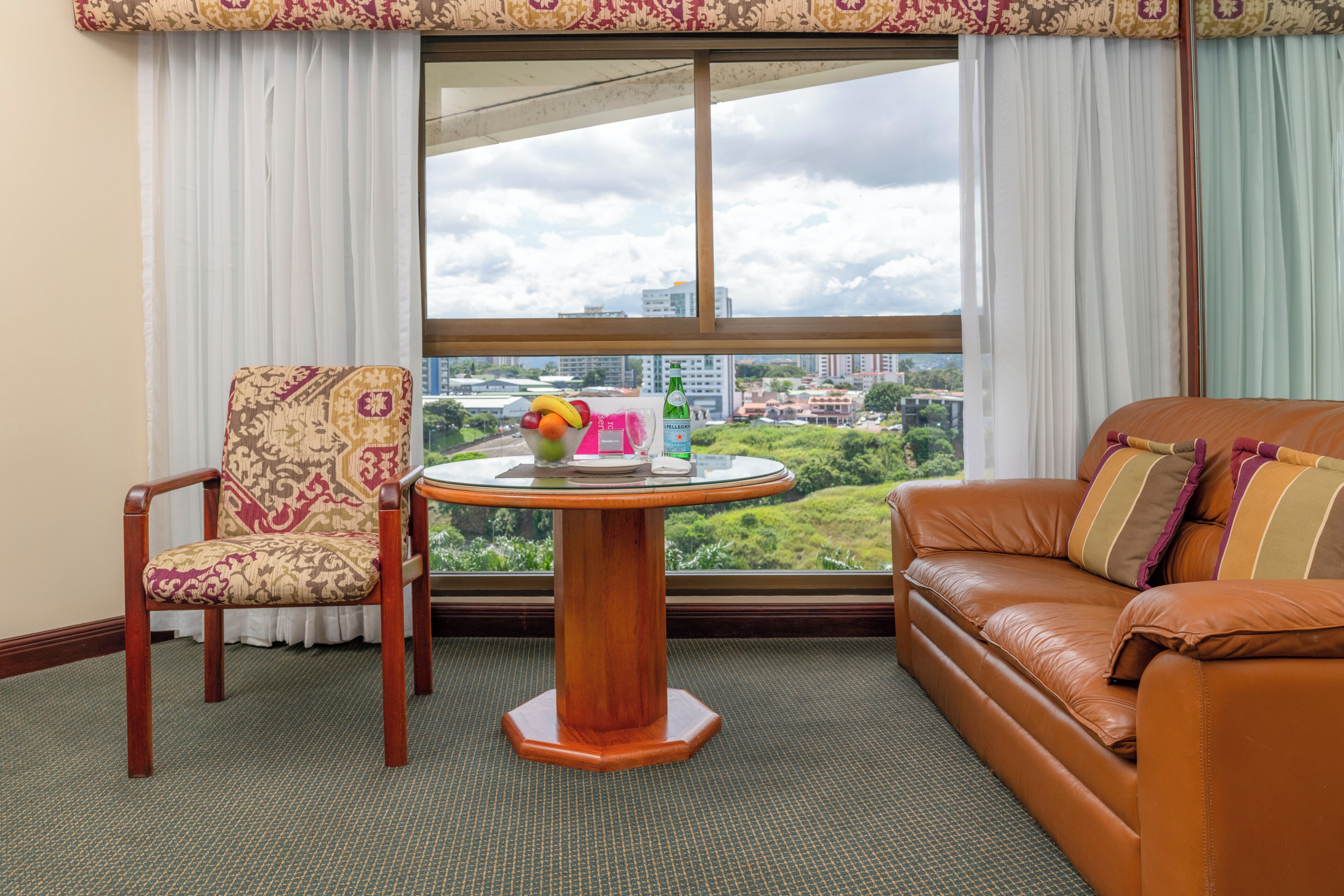 Quarto com vista para a cidade, uma mesa de madeira redonda com uma cesta de frutas e uma garrafa, uma poltrona estampada e um sofá de couro com almofadas listradas.