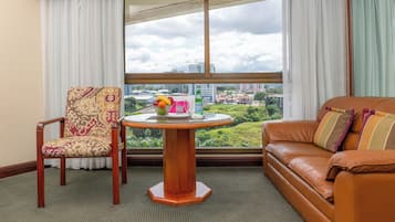 Quarto com vista para a cidade, uma mesa de madeira redonda com uma cesta de frutas e uma garrafa, uma poltrona estampada e um sofá de couro com almofadas listradas.