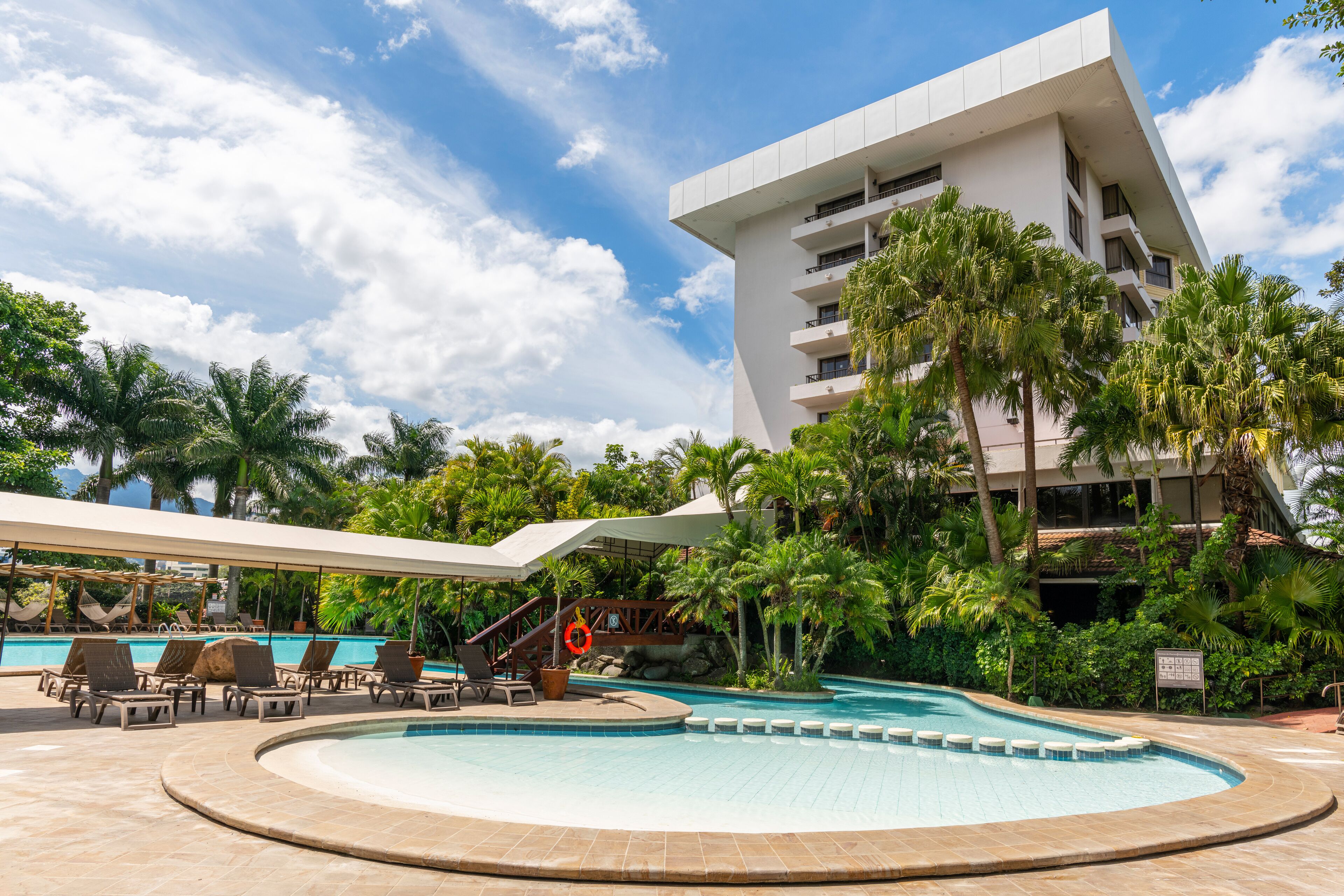 A modern hotel with a pool area, palm trees, and a clear sky.