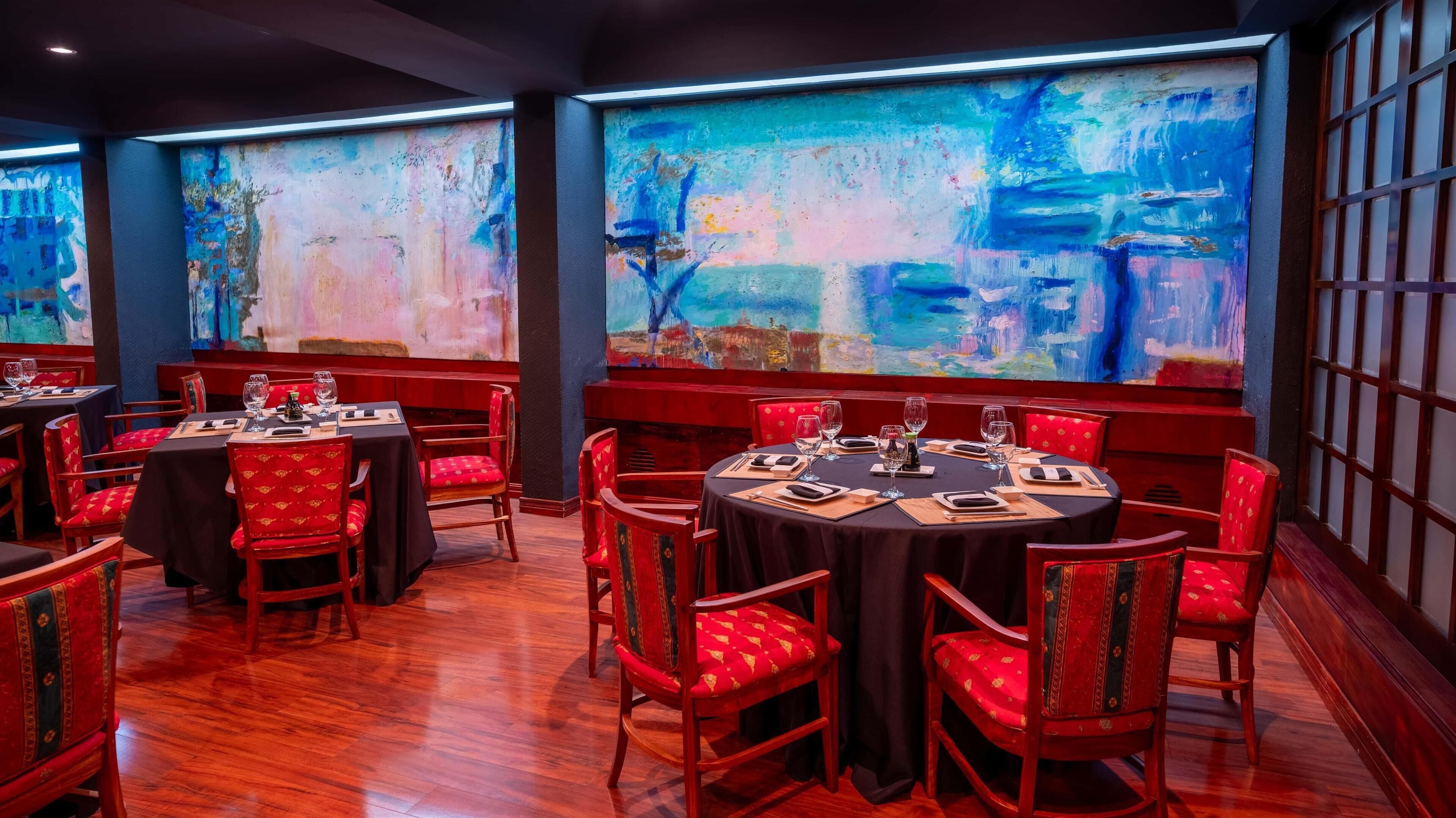 A restaurant with round tables and red upholstered chairs, large abstract paintings on the walls, and wooden flooring.