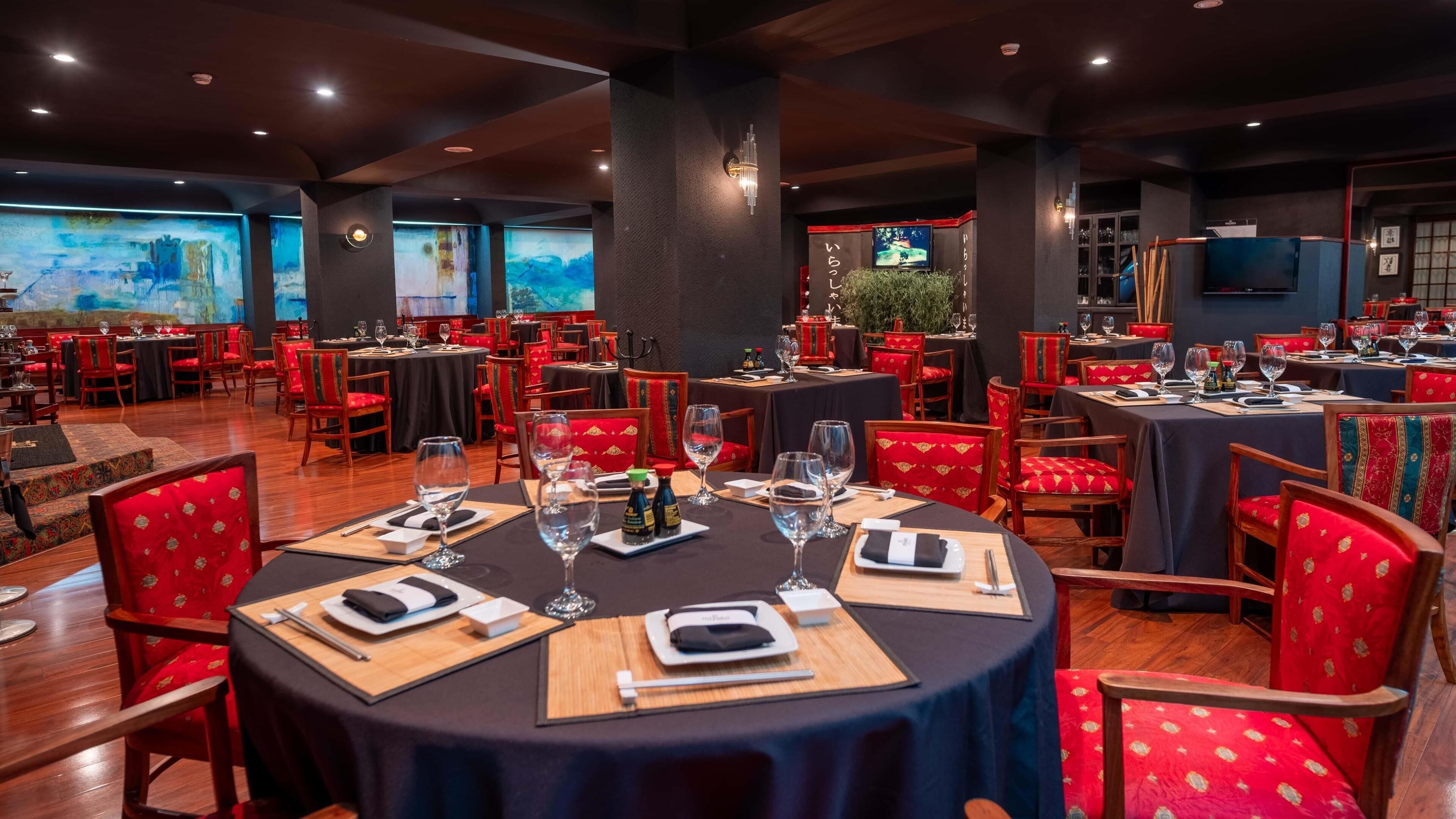 A restaurant with round tables and red chairs, featuring a large mural on the wall and a television screen.