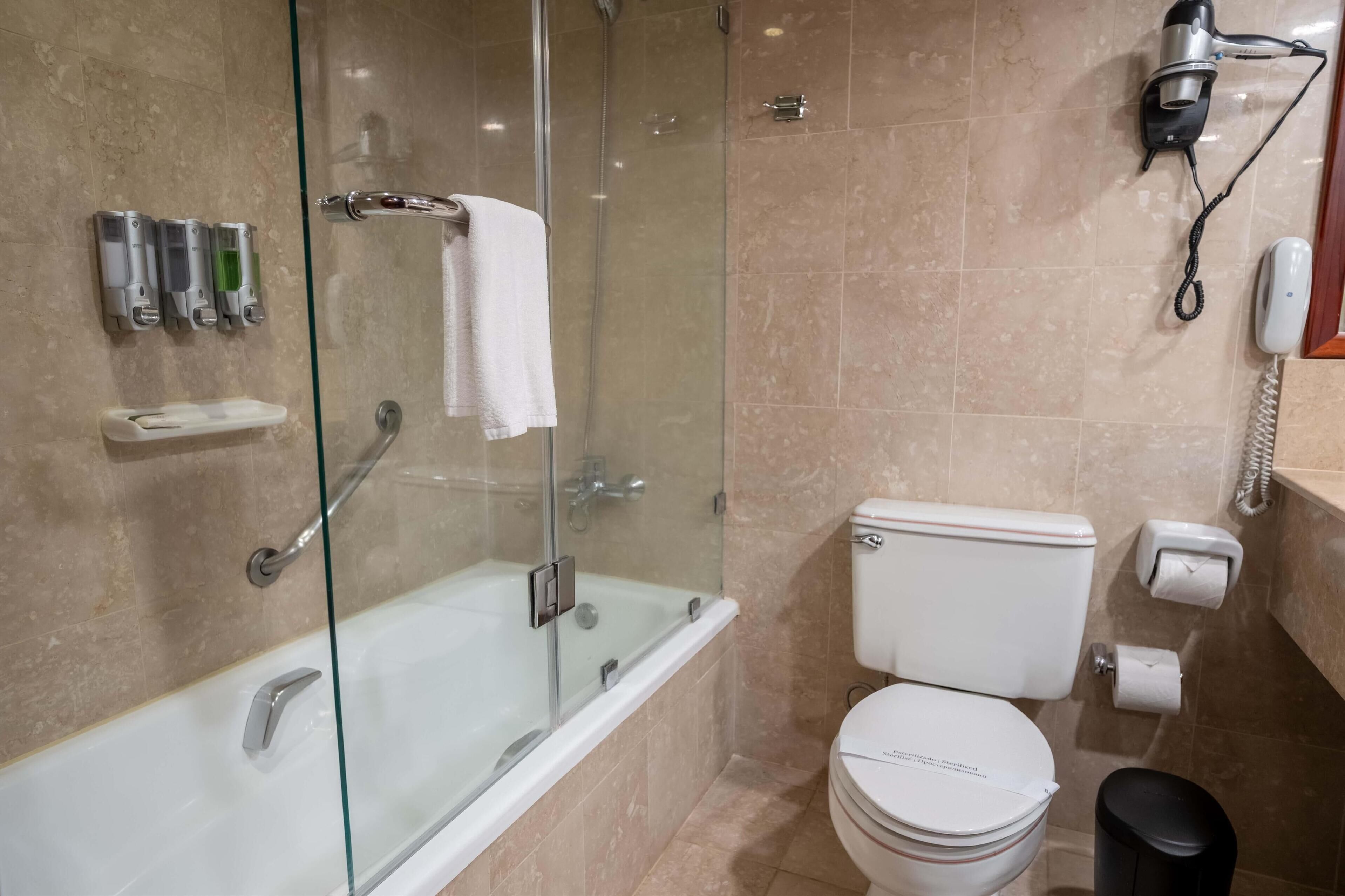 Un baño moderno con ducha de vidrio, bañera, inodoro y secador de pelo montado en la pared.