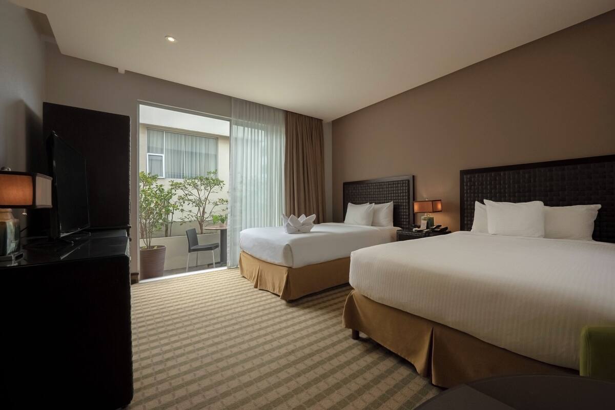 A hotel room with two beds, a desk, a TV, and a balcony with plants.
