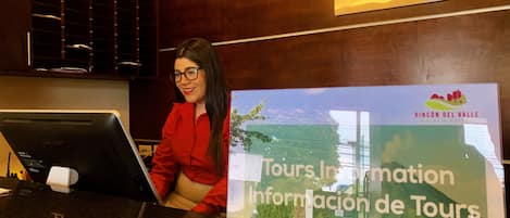 A reception desk with a digital screen displaying tour information, a wooden reception area, and a sign indicating the hotel name.