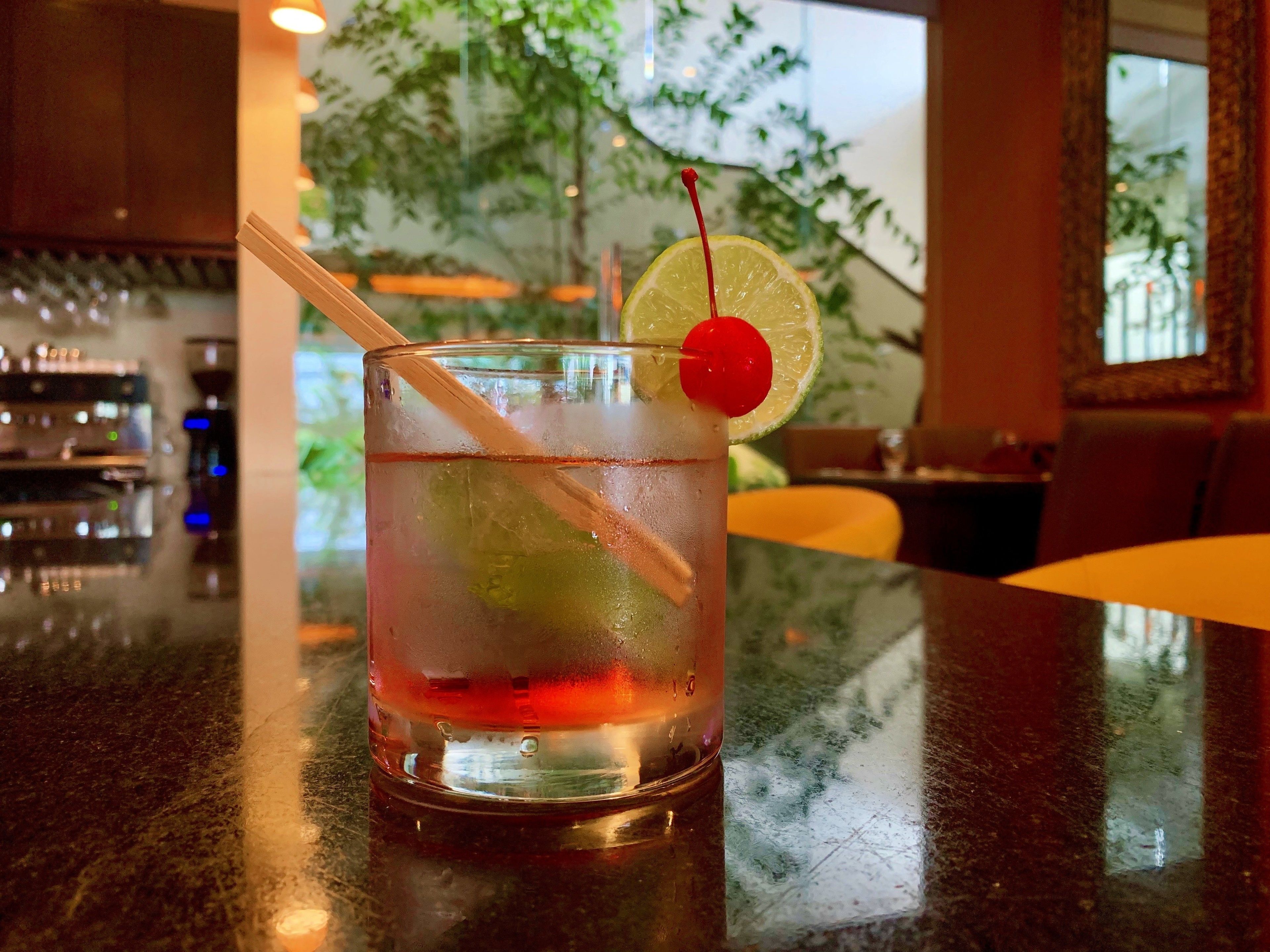A glass of iced beverage with a straw and a cherry on a slice of lime, placed on a bar counter.