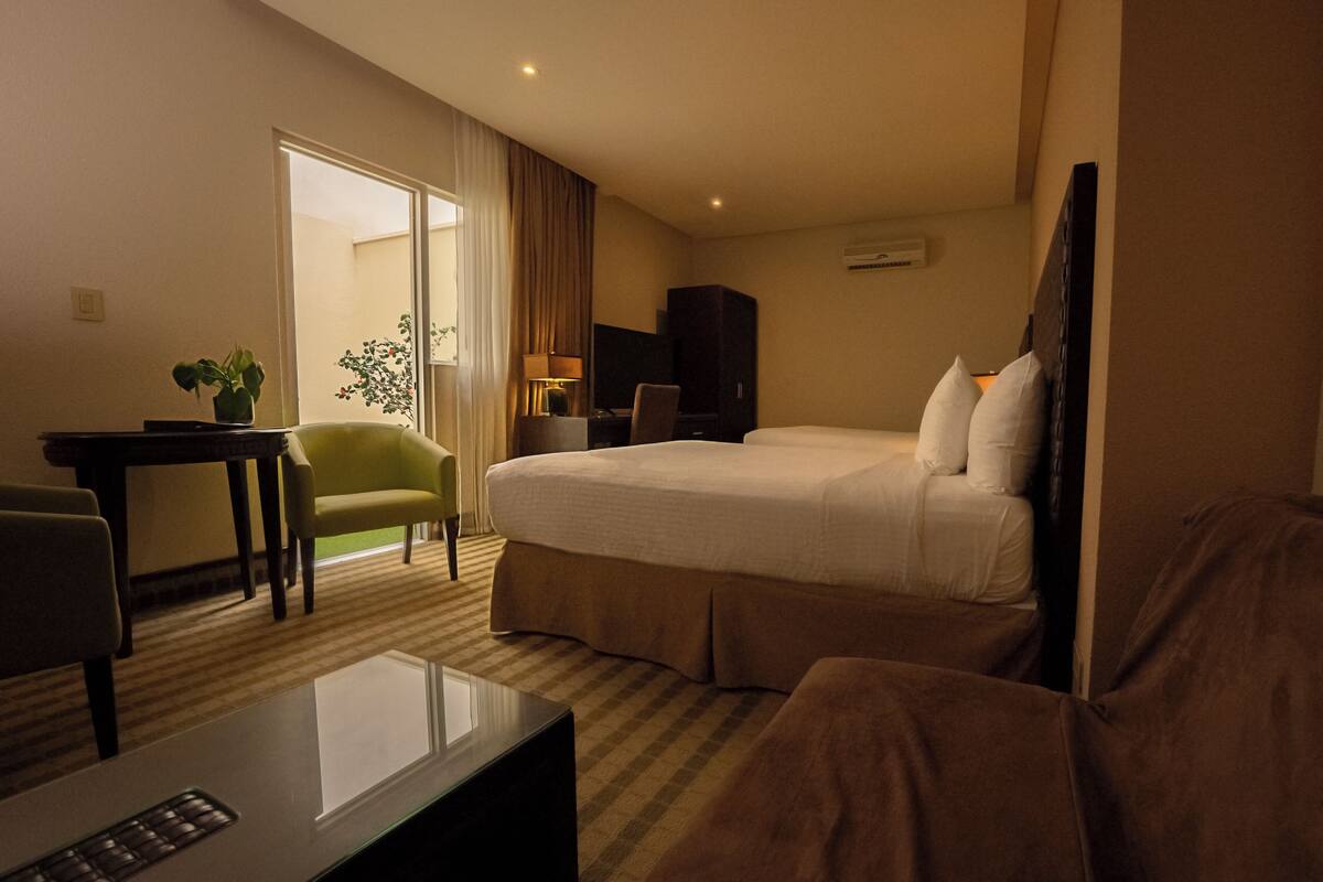 A hotel room with a bed, a desk, a chair, a TV, and a plant.