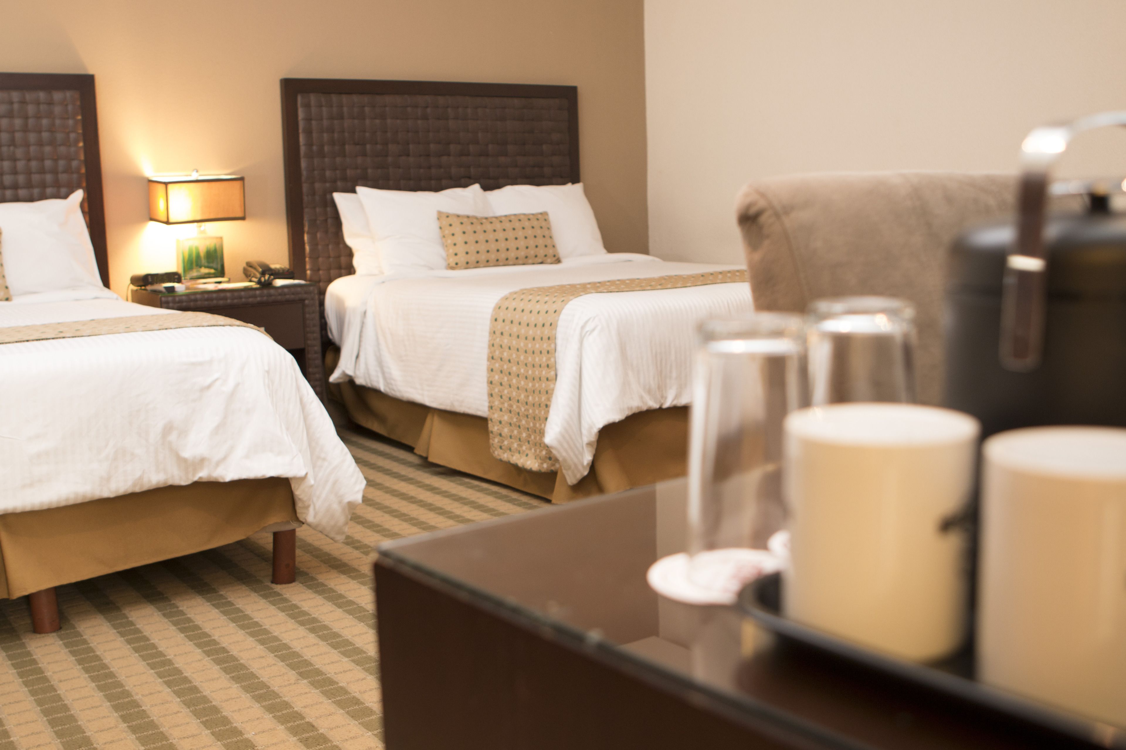 A hotel room with two beds, a nightstand with a lamp, and a coffee table with mugs.