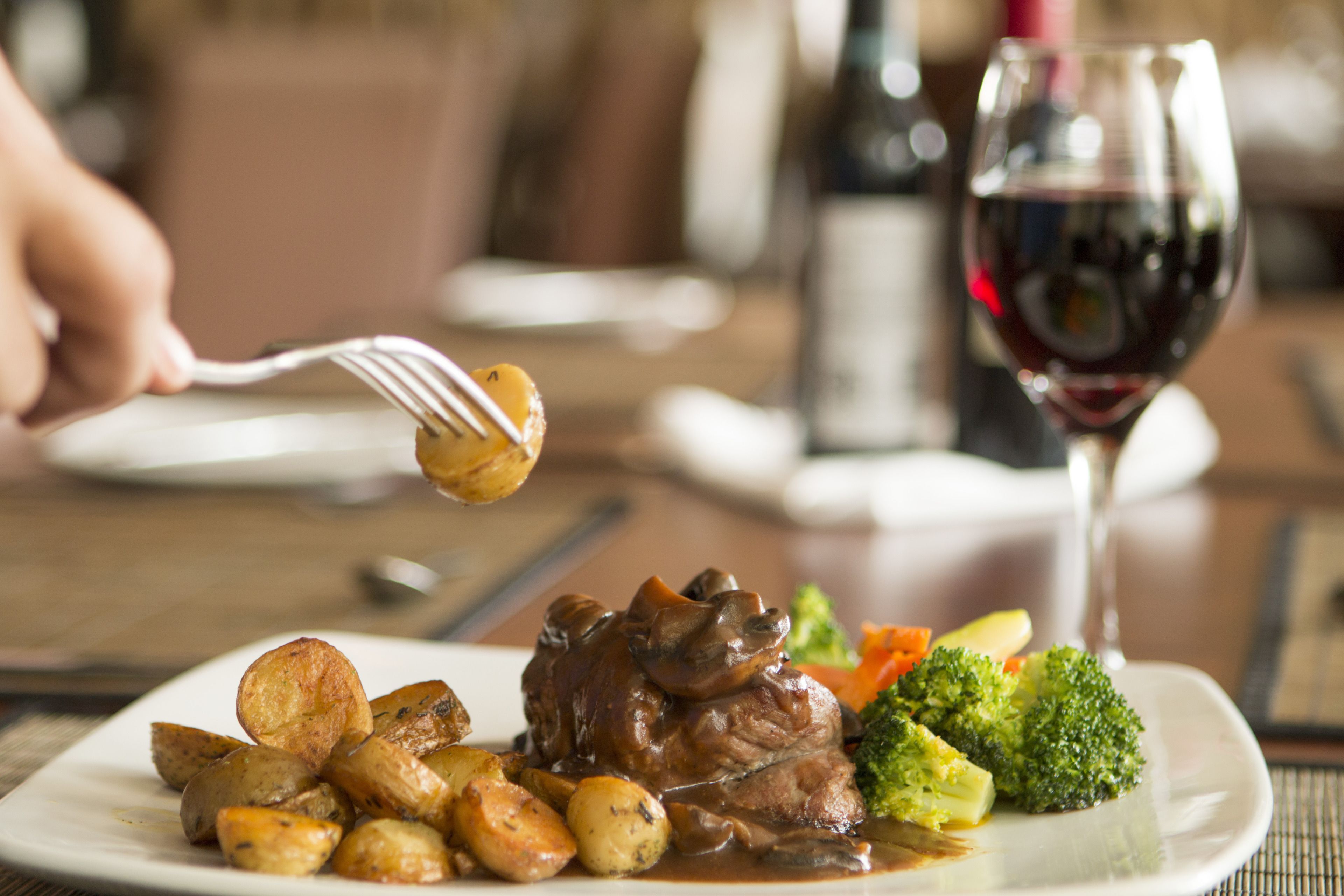 A plate of food with a fork lifting a piece of meat, a glass of red wine, and a bottle of wine in the background.