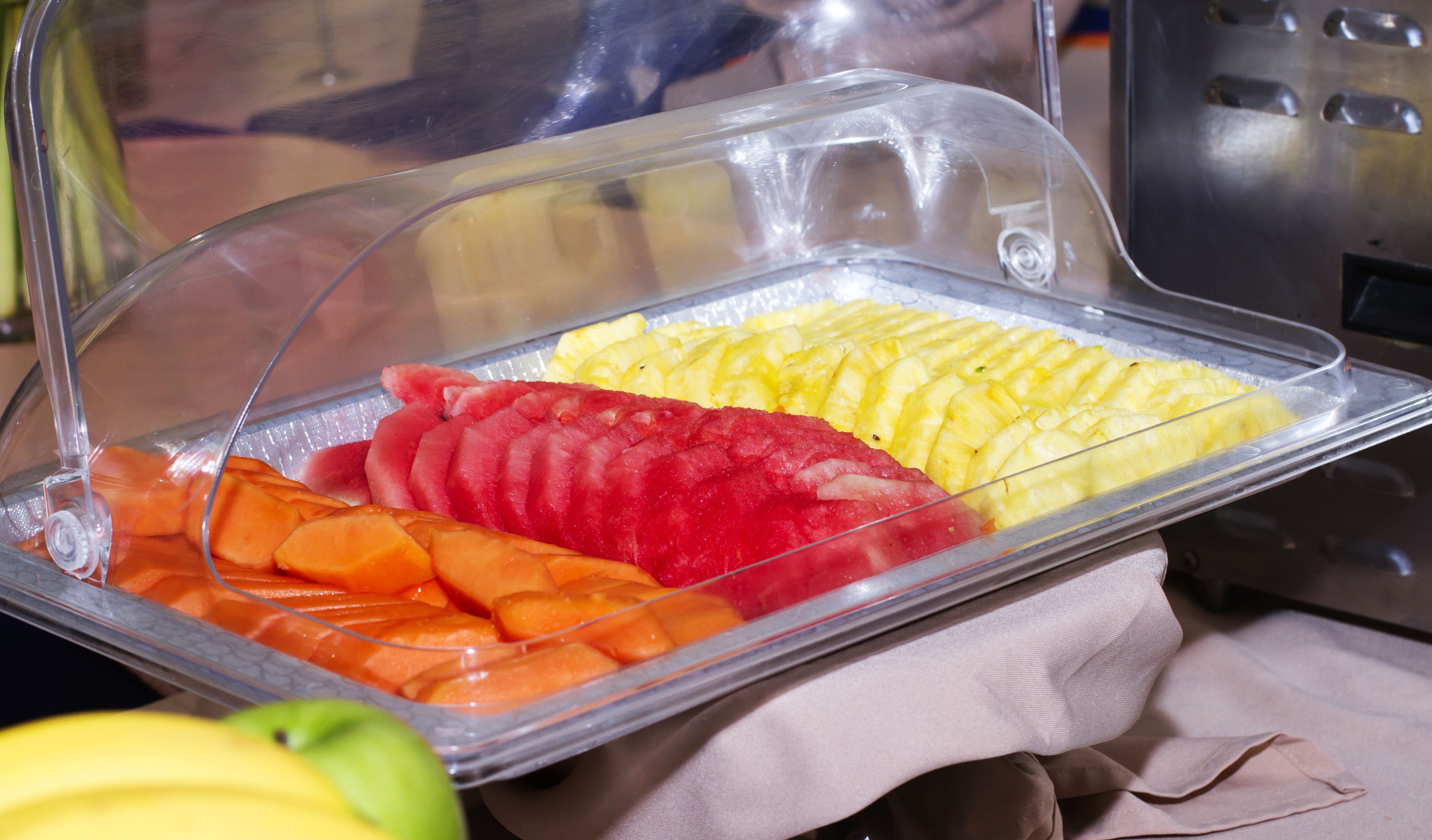 Un bufé con frutas y verduras cortadas bajo una cubierta de plástico transparente.
