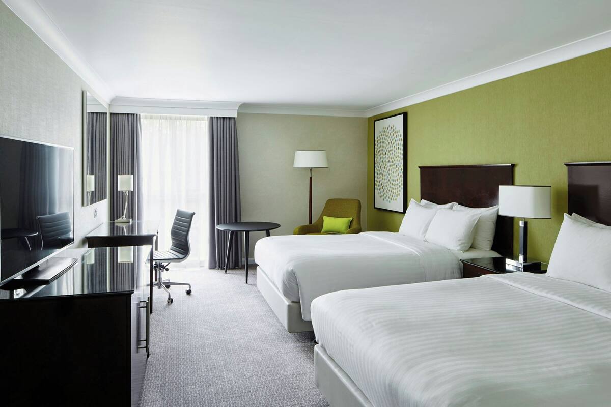A hotel room with two beds, a desk, a chair, a TV, and a lamp.