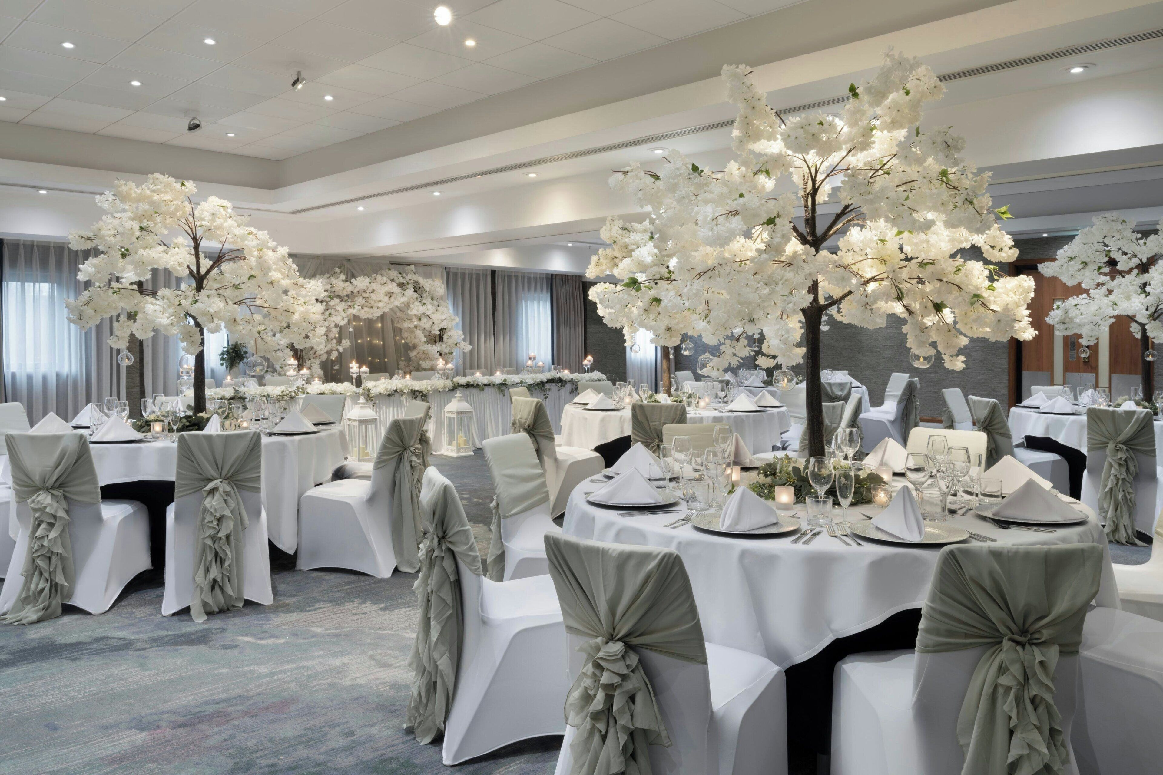 En festlokal med runda bord uppdukade för ett formellt evenemang, dekorerade med vita blomsterarrangemang och eleganta bordsdekorationer.