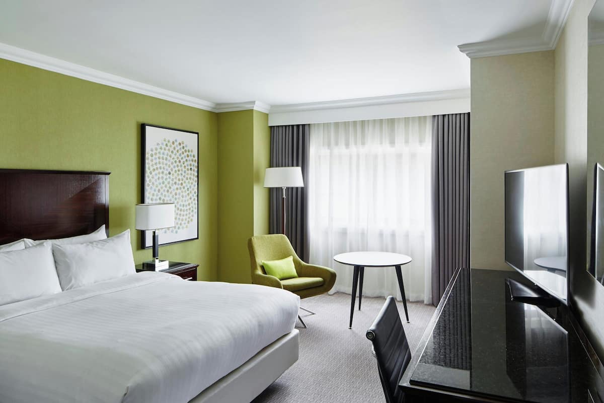 A hotel room with a large bed, a desk with a chair, a television, a lamp, and a window with curtains.