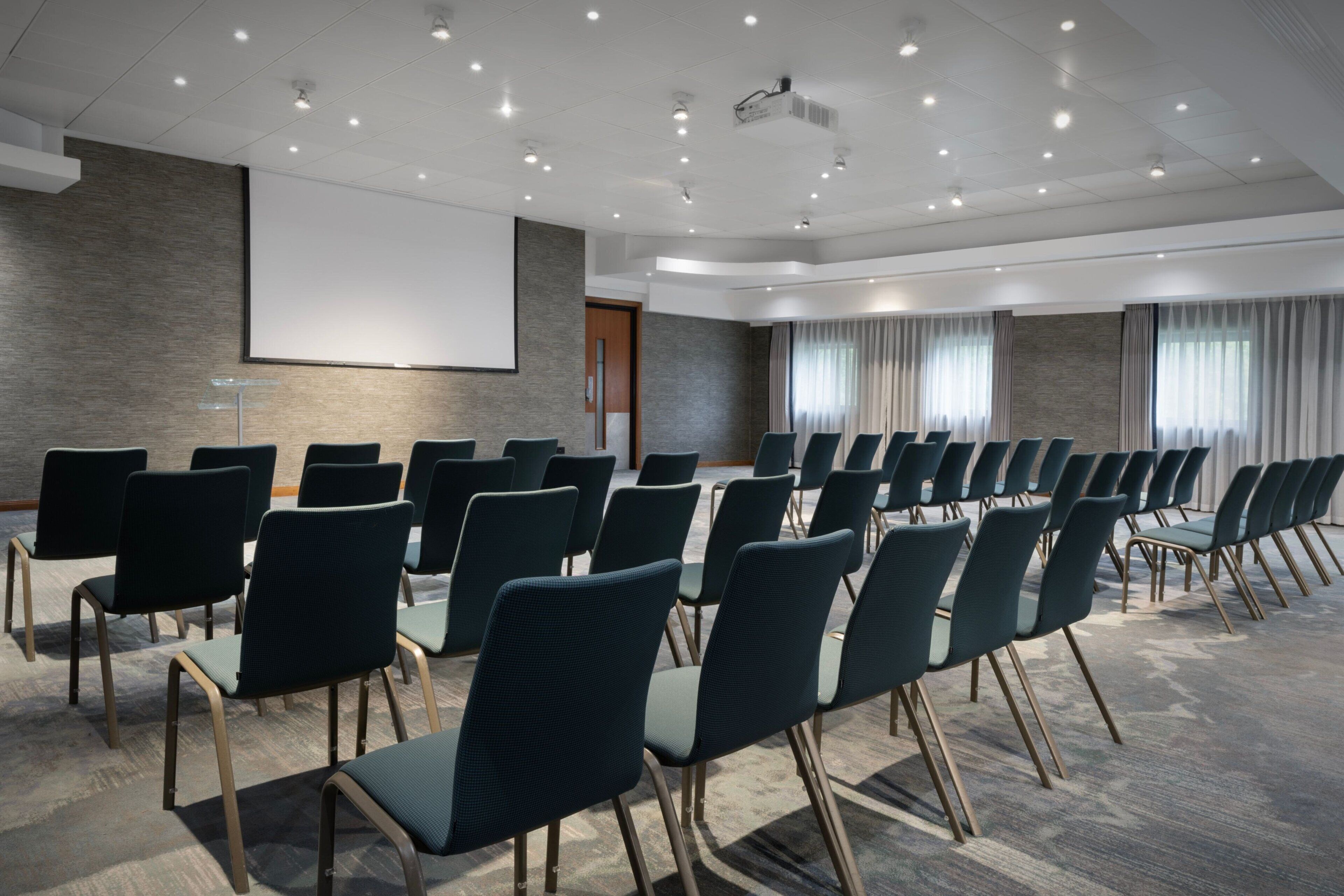 Ein Konferenzraum mit Reihen von Stühlen, einer Projektionsleinwand und einem großen Fenster mit Vorhängen.