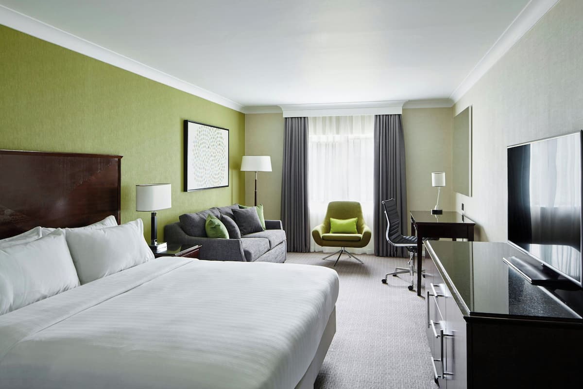 A hotel room with a large bed, a desk with a TV, a green armchair, and a sofa with cushions.