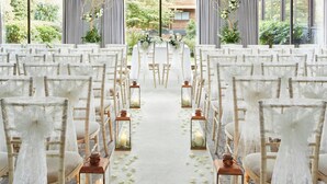 Une allée décorée pour un mariage, avec des chaises blanches et des lanternes, menant à une table ornée de compositions florales.