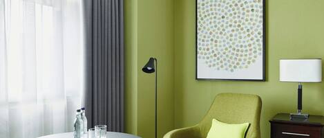 Una habitación con una mesa redonda, un sillón verde, una lámpara de pie, una mesita de noche con lámpara, una obra de arte abstracta enmarcada y una ventana con cortinas.
