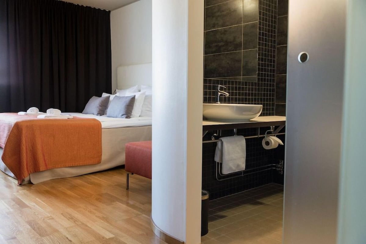 A modern hotel room with a bed, a bedside table, a small stool, a bathroom with a sink and mirror, and a dark curtain.