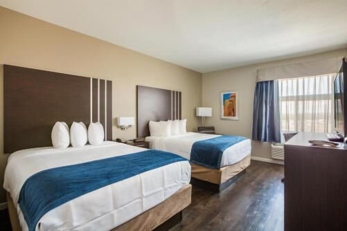Une chambre d’hôtel avec deux lits, un bureau, une chaise, une lampe et une fenêtre avec des rideaux.