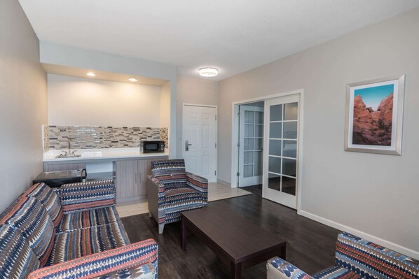 A modern living room with patterned sofas, a wooden coffee table, and a kitchen area with a microwave and tiled backsplash.