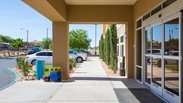 A covered outdoor area with a sidewalk leading to a building entrance, potted plants, and parked cars.