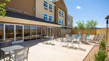 A poolside area with lounge chairs and tables, a pool, and a building with large windows and a wooden fence.
