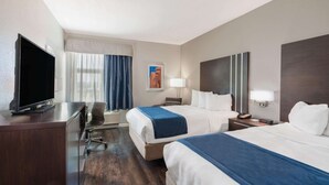 Ein Hotelzimmer mit zwei Betten, einem Schreibtisch mit Fernseher, einem Sessel, einer Lampe und einem Bild an der Wand.