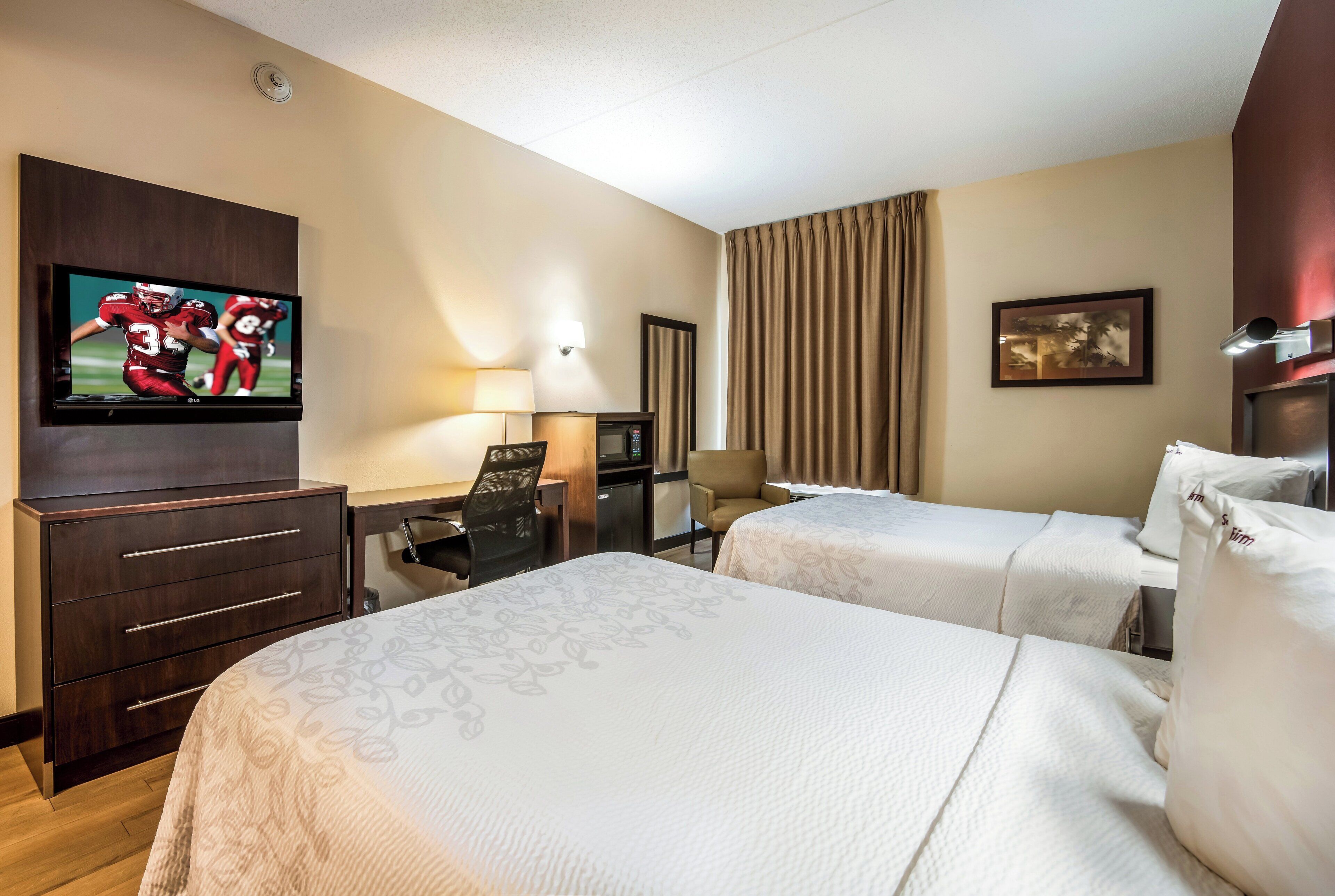 A hotel room with two beds, a TV, a desk with a chair, a lamp, a microwave, and a painting on the wall.