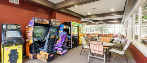 A gaming area with arcade machines, a seating area with tables and chairs, and large windows providing natural light.