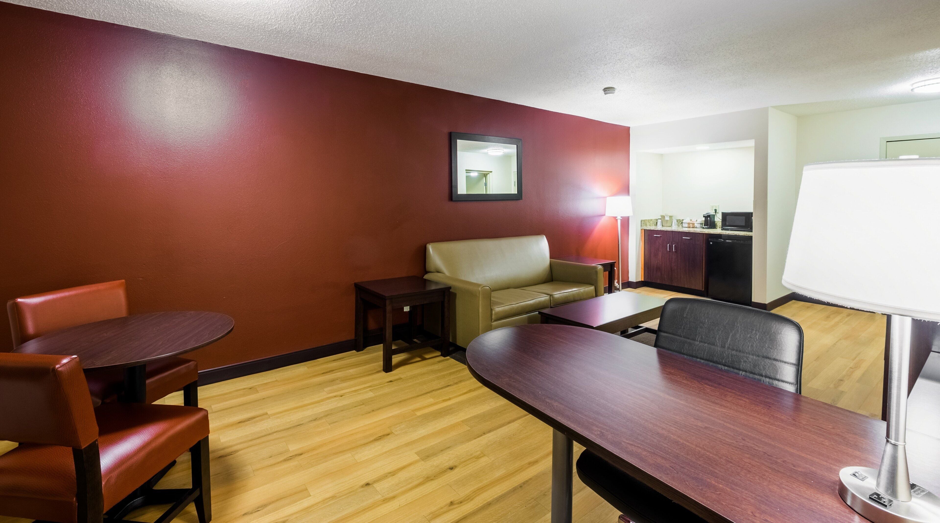 A hotel room with a dining table, two chairs, a sofa, a small table, a lamp, and a kitchenette.