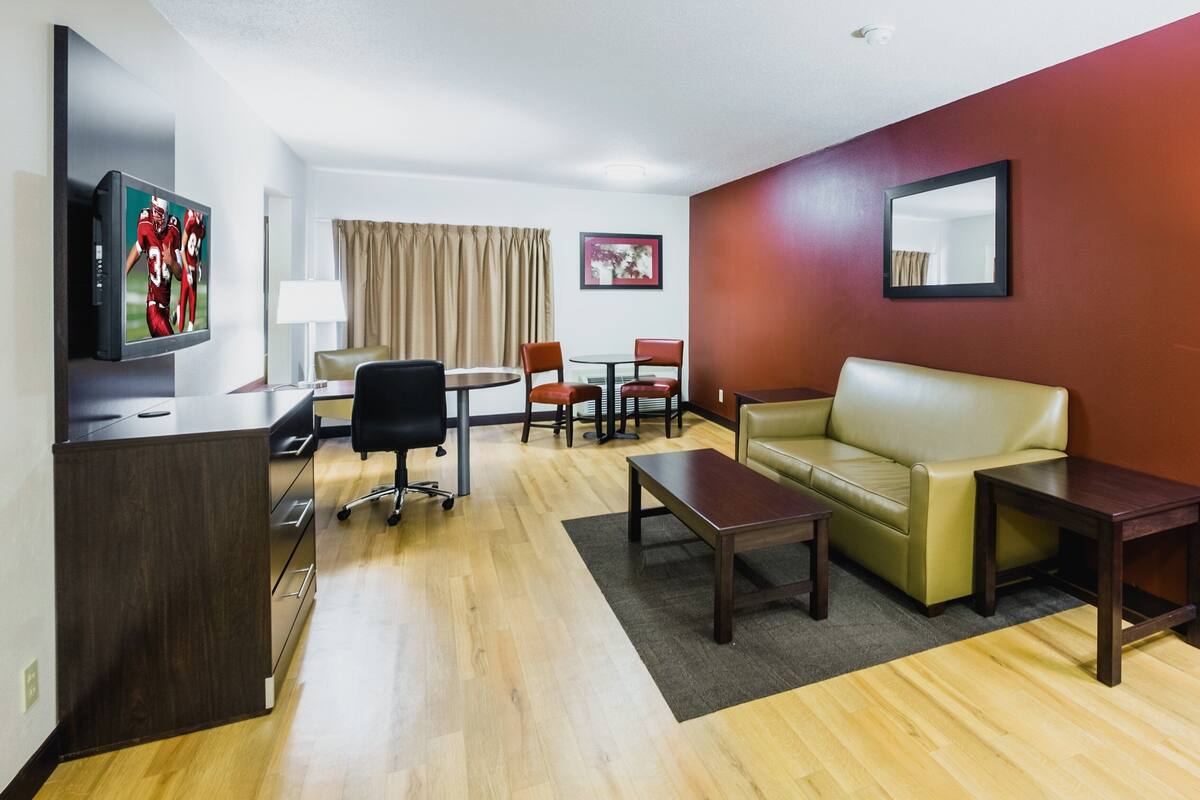Habitación de hotel con piso de madera, sofá de cuero, escritorio con televisor, mesa de comedor con sillas y espejo en la pared.