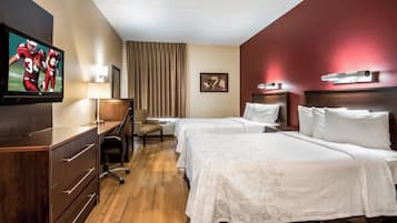 A hotel room with a TV, desk, chair, two beds, and a window with curtains.