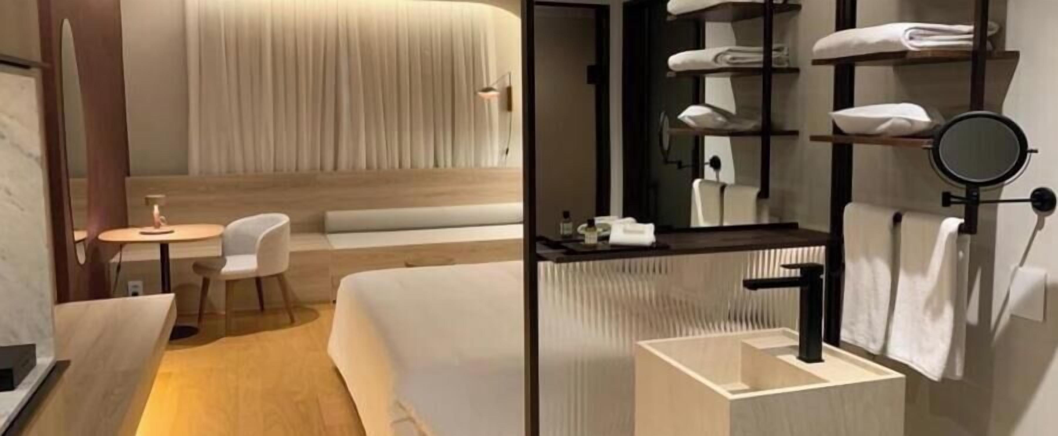 Une chambre d’hôtel moderne équipée d’un lit, d’un petit coin salon, d’un bureau, d’un miroir rond et d’un lavabo intégré.