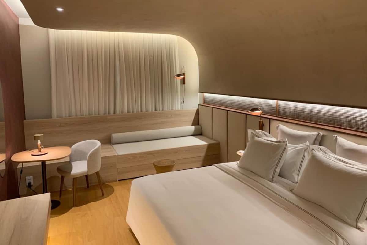 Ein modernes Hotelzimmer mit einem großen Bett, einer Sitzecke mit einem kleinen Tisch und einem großen Fenster mit Vorhängen.