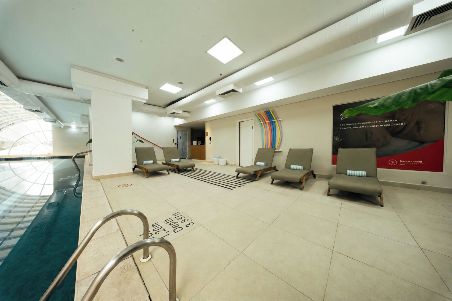 Um hall de hotel com piscina, cadeiras de descanso, escadaria e um cartaz na parede.