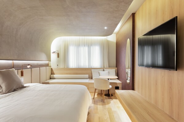 Una habitación de hotel moderna con una cama grande, un escritorio, una silla, un televisor de pantalla plana montado en la pared y un ventanal con cortinas.