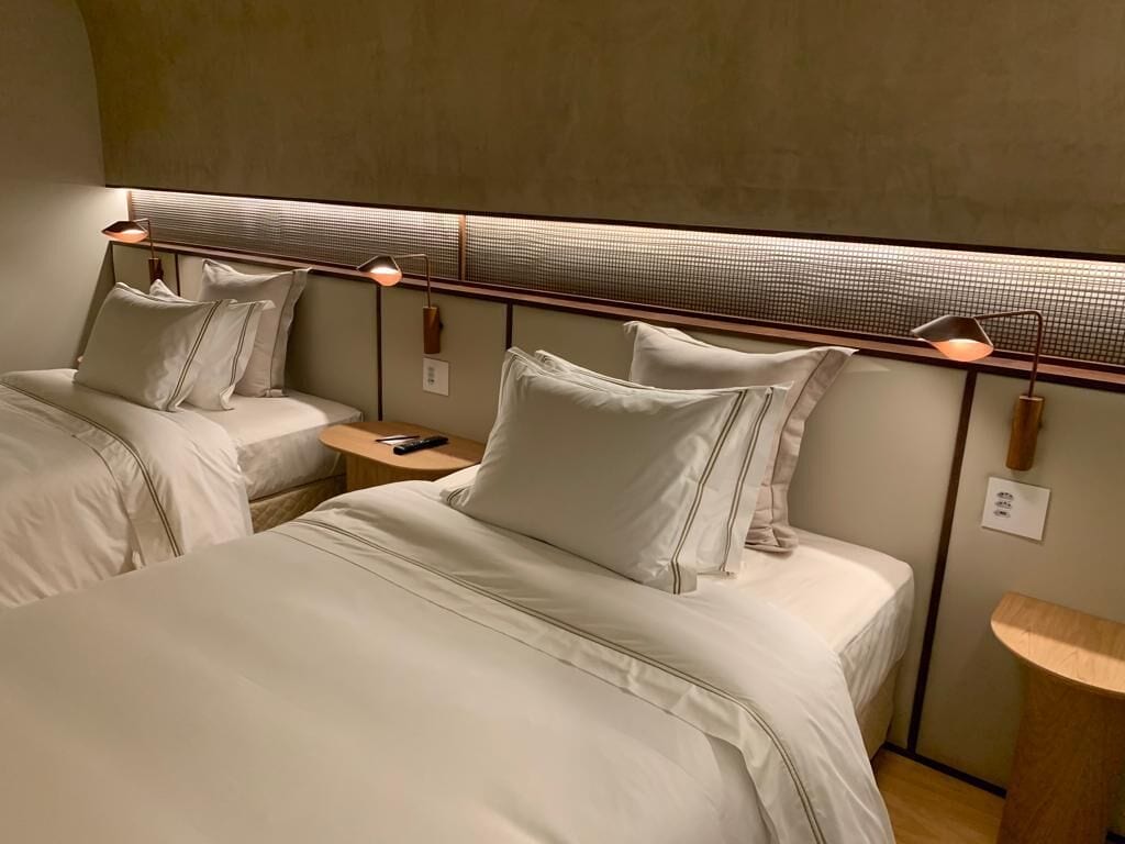 Quarto de hotel com duas camas, cabeceira com iluminação integrada e uma pequena mesa de cabeceira de madeira.