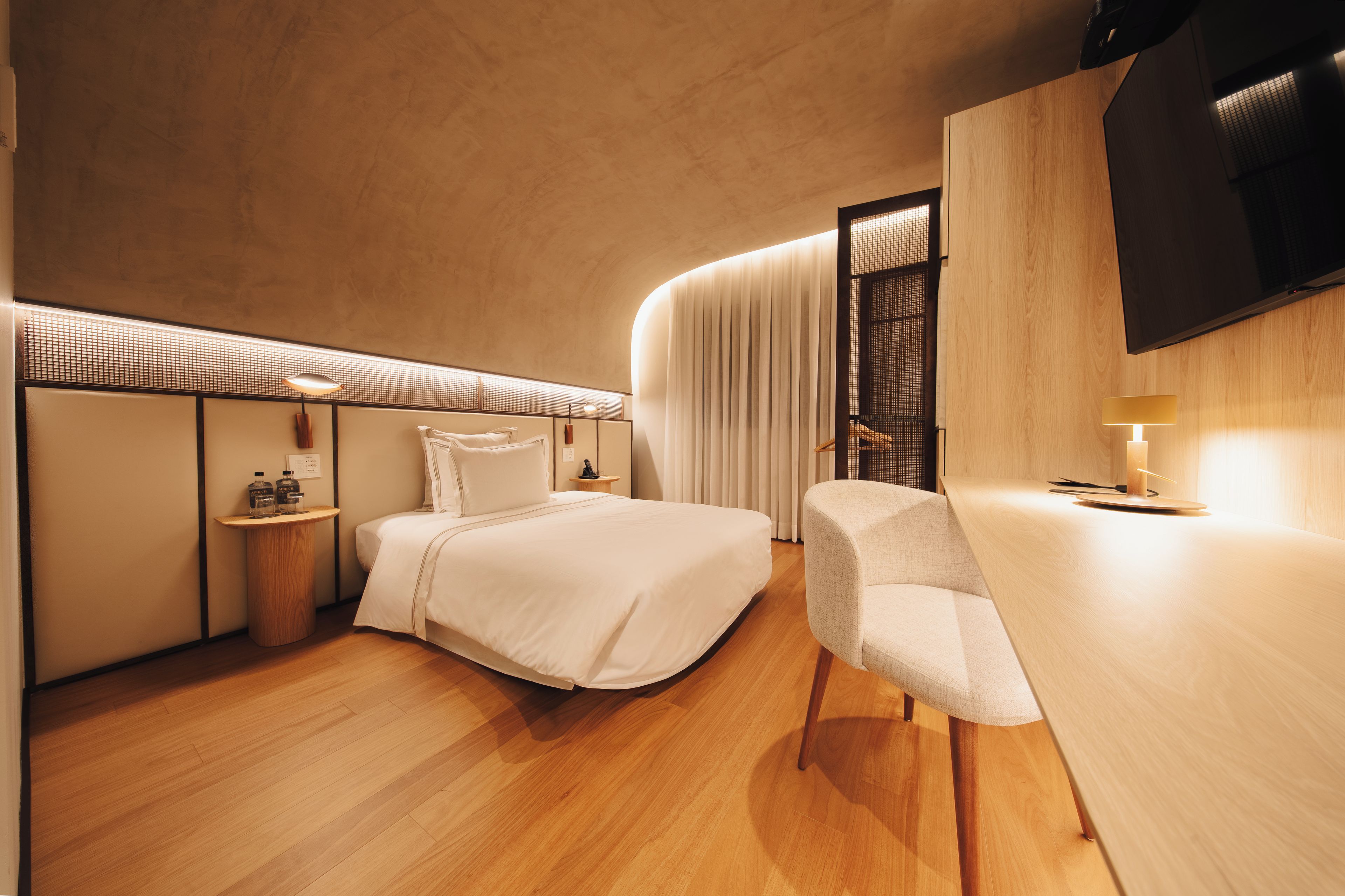 A modern hotel room with a bed, a desk, a chair, a TV, and a minibar.