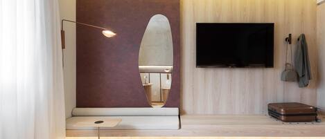 A modern hotel room with a large bed, a flat-screen TV mounted on the wall, a seating area with a bench, and a suitcase on the floor.