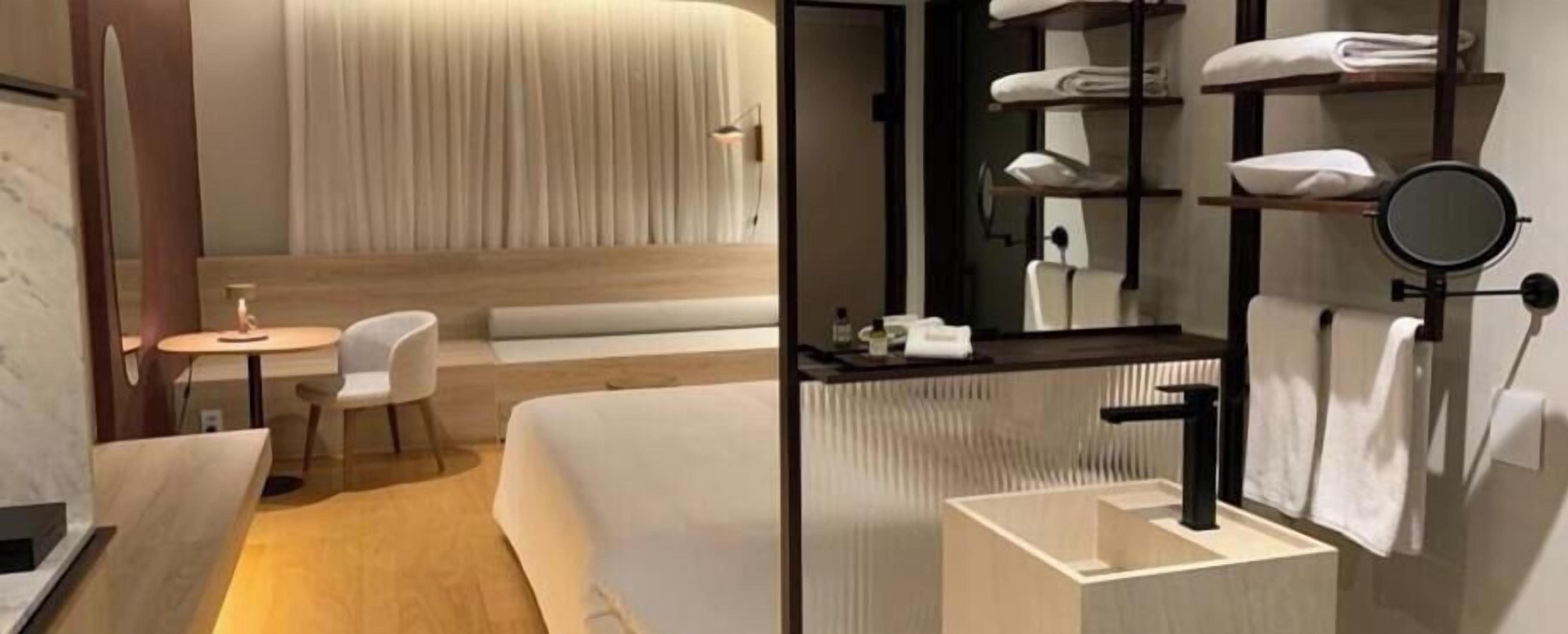 Une chambre d’hôtel moderne équipée d’un lit, d’un petit coin salon, d’un bureau, d’un miroir rond et d’un lavabo intégré.