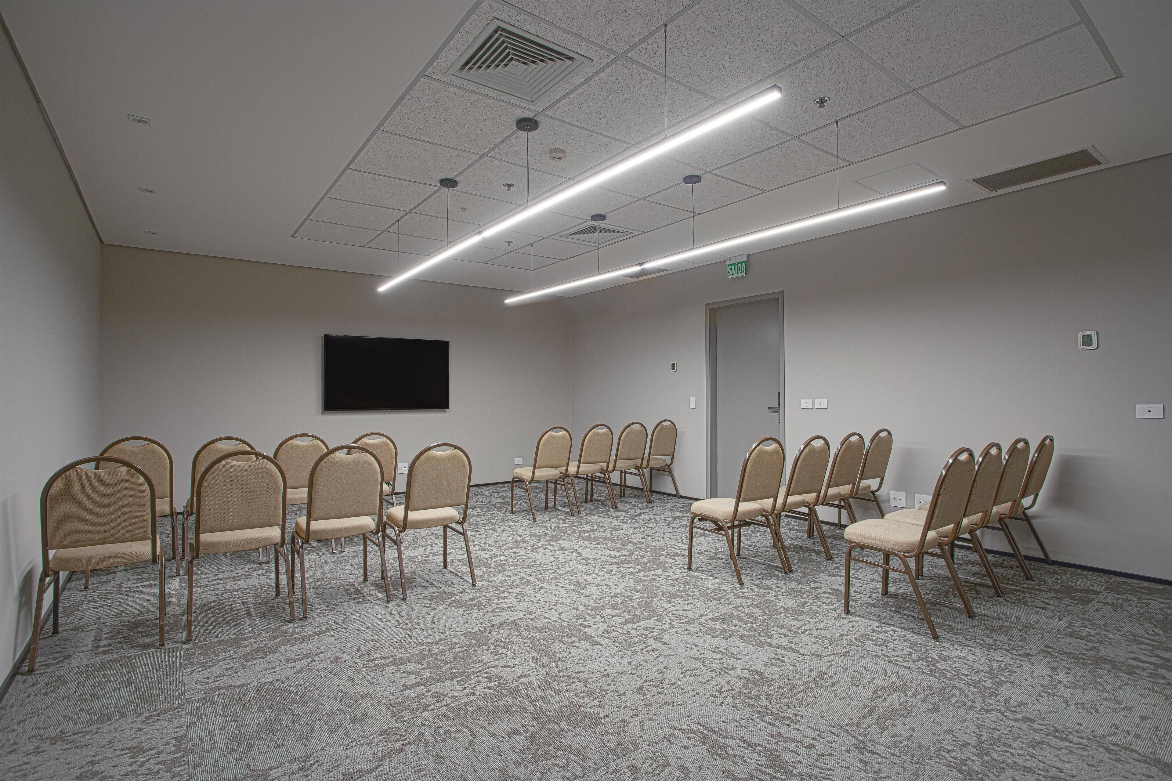 A conference room with beige chairs arranged in a semi-circle, a flat-screen TV mounted on the wall, and a carpeted floor.