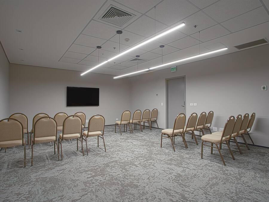 A conference room with beige chairs arranged in a semi-circle, a flat-screen TV mounted on the wall, and a carpeted floor.